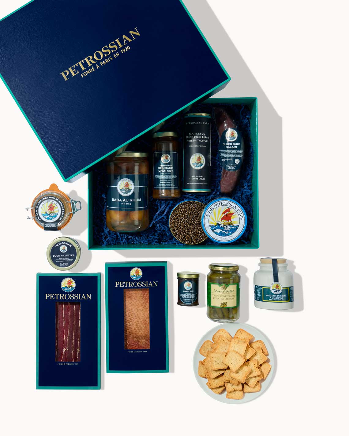 An overhead image of a navy-Blue Petrossian gift box displaying Petrossian delicacies including duck prosciutto, smoked duck breast, caviar and cornichons  on a white background.