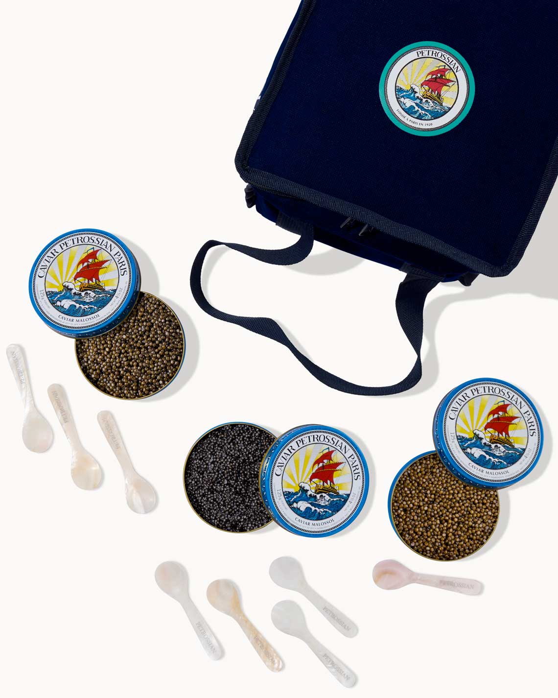 an overhead image of a navy-Blue Petrossian tote bag with three tins of different caviars and many pearl spoons laid out on a white background.
