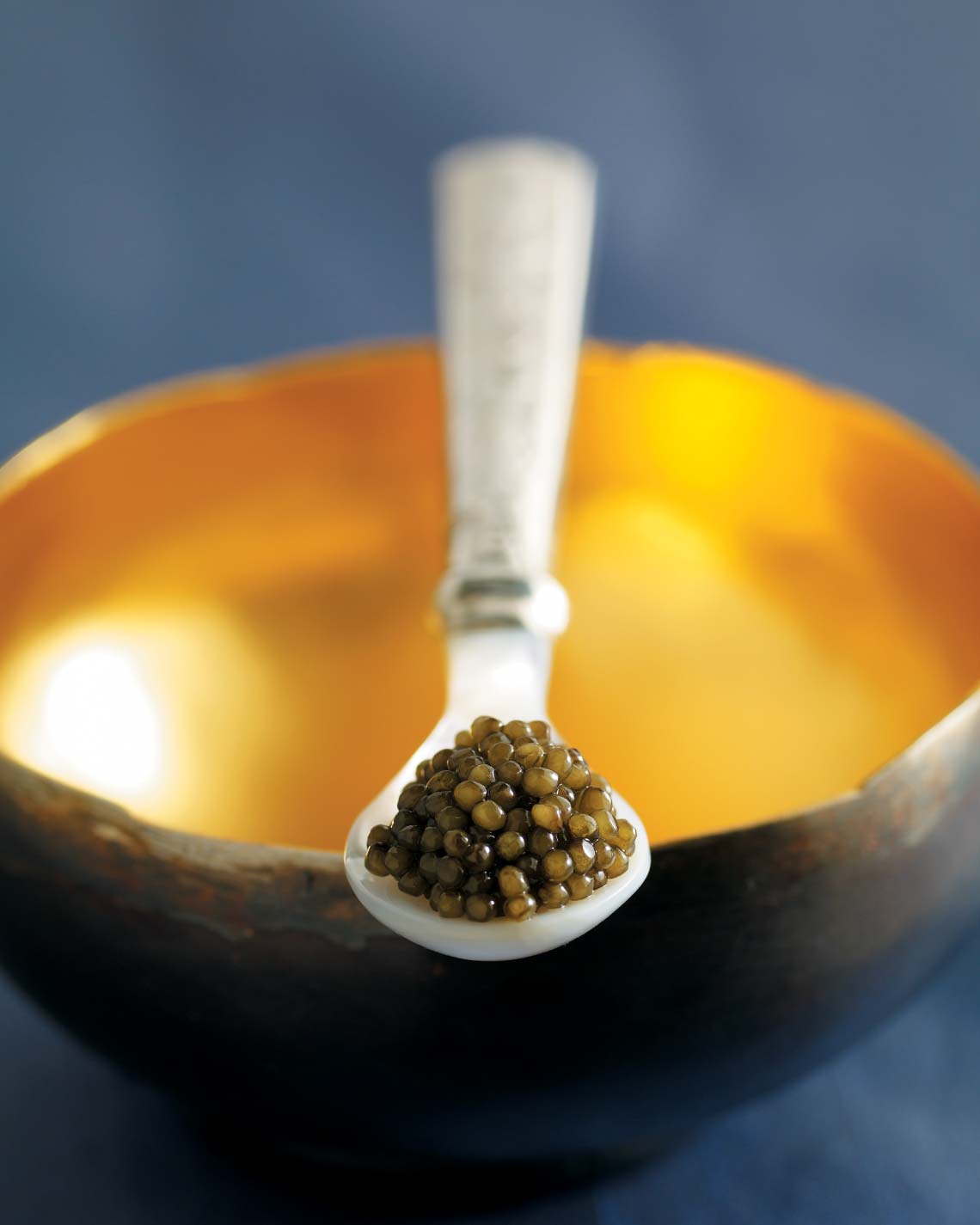 A tasteful dollop of light green Ossetra caviar sits elegantly on an ornate pearl spoon that sits across a brown ceramic bowl with a bright gold interior.