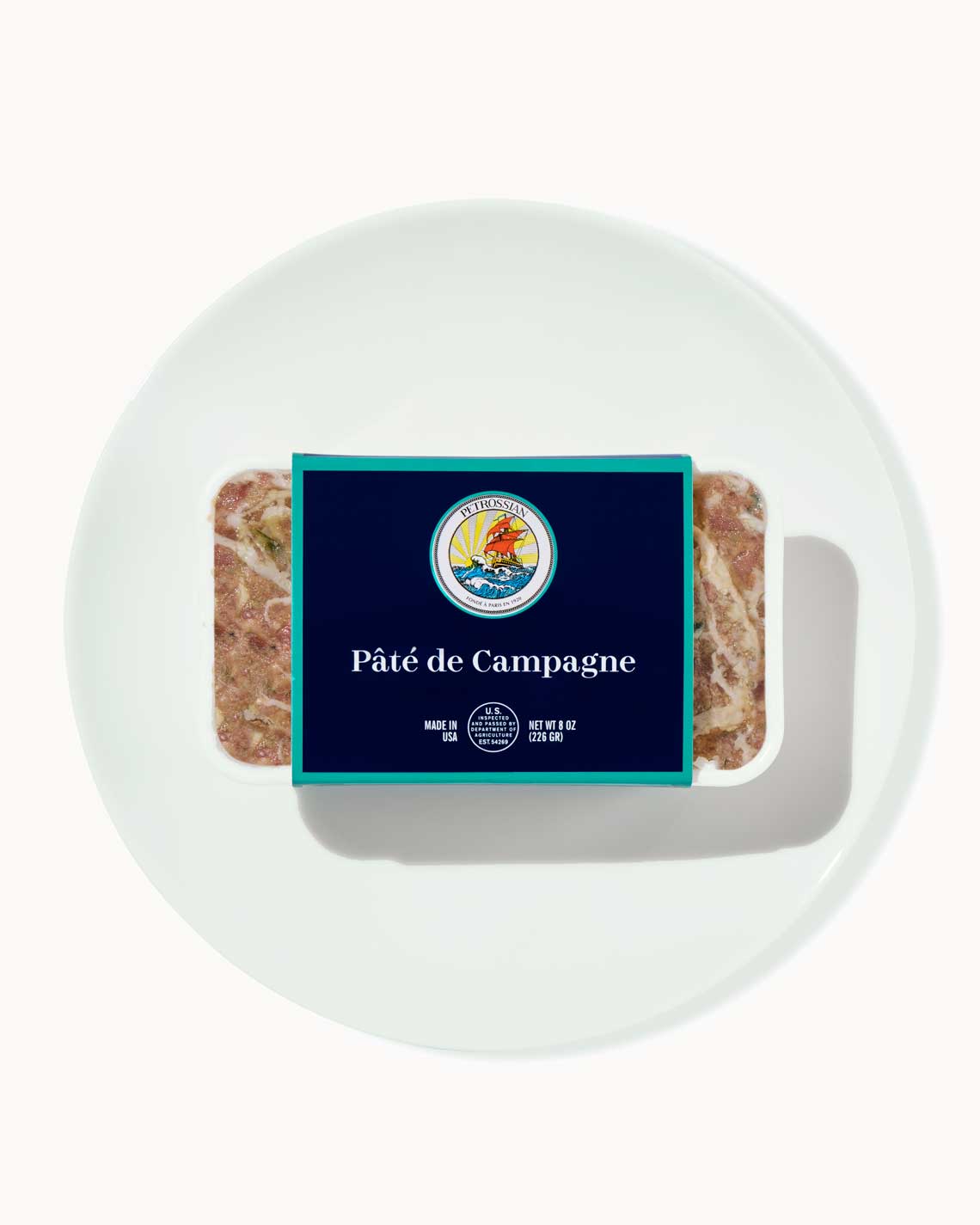 a terrine of rich, succulent pâté sits on a white plate on a white background. the terrine is wrapped in a navy blue and teal Petrossian label.