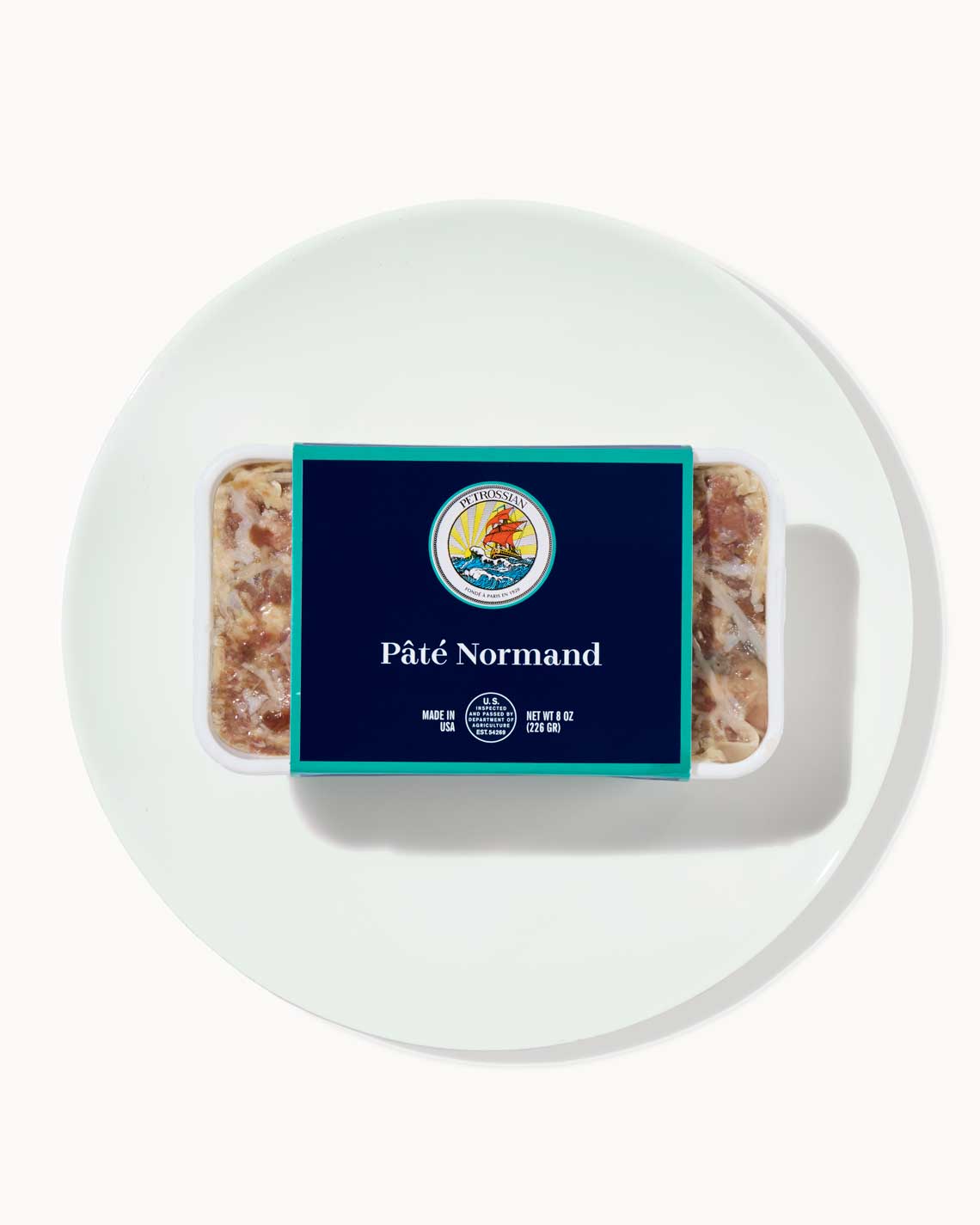 a terrine of rich, succulent pâté sits on a white plate on a white background. the terrine is wrapped in a navy blue and teal Petrossian label.