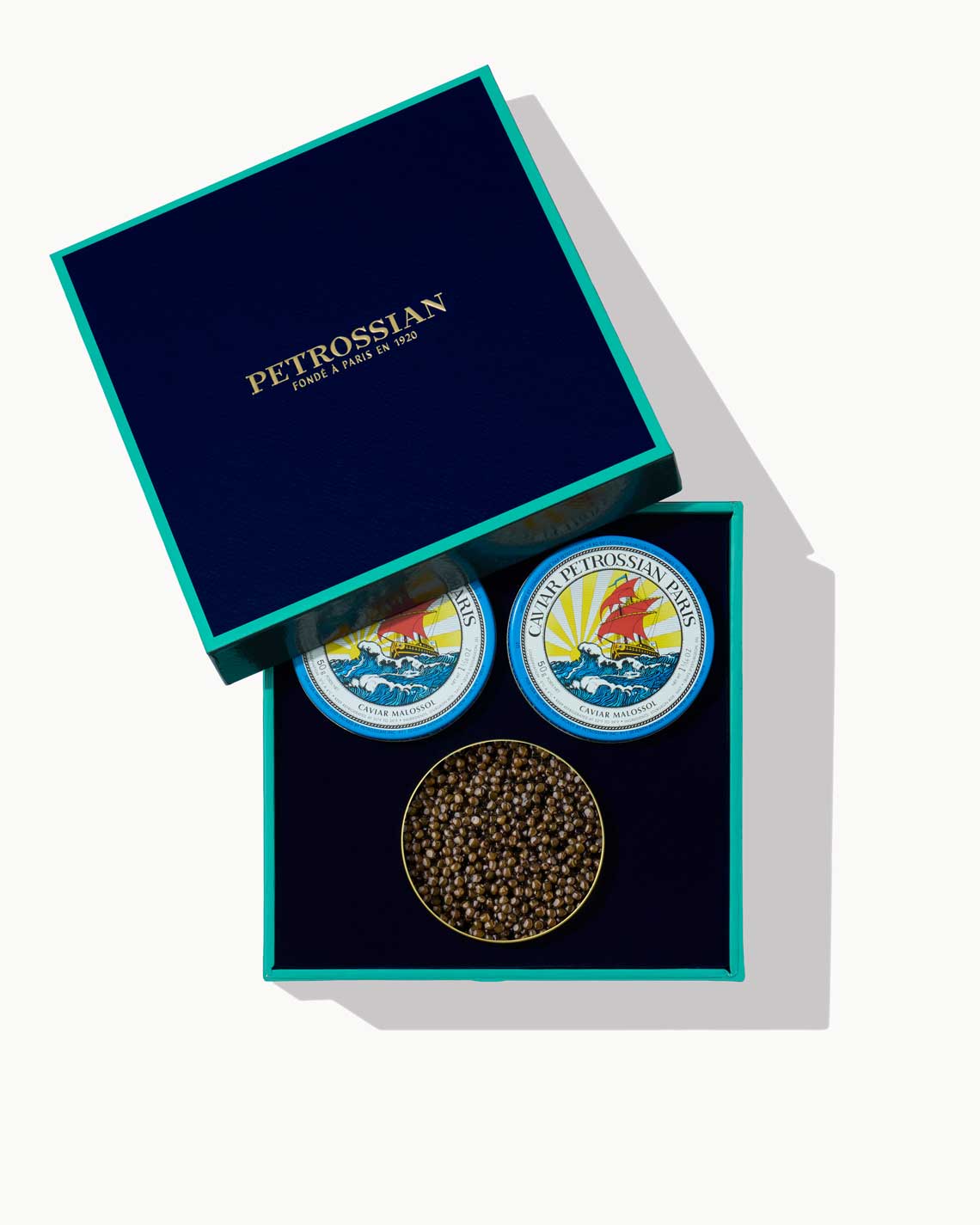 a navy blue Petrossian gift box with teal trim, holding threee tins of Petrossian caviar, one opened, and sitting on a white background.