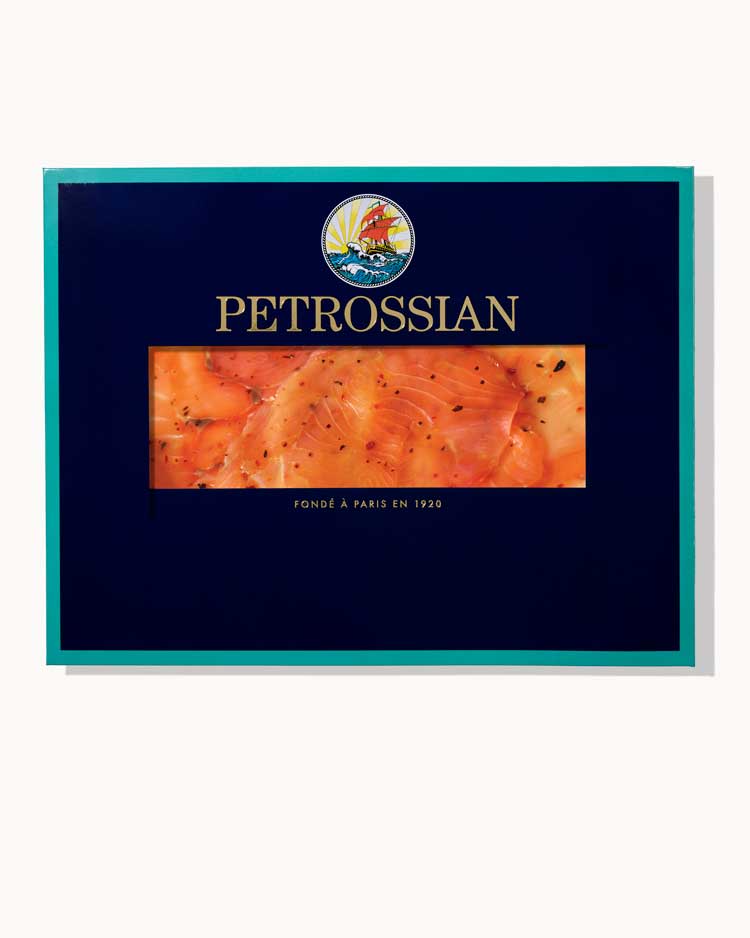 A navy blue Petrossian box with teal trim on a white background. A juicy filet of rich Smoked Salmon carpaccio can be seen through a cut-out in the center of the box.