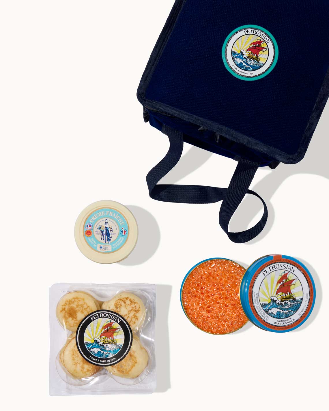 an overhead image of a navy-Blue Petrossian tote bag with a tin a bright red salmon roe, blinis and a jar of creme fraiche laid out on a white background.
