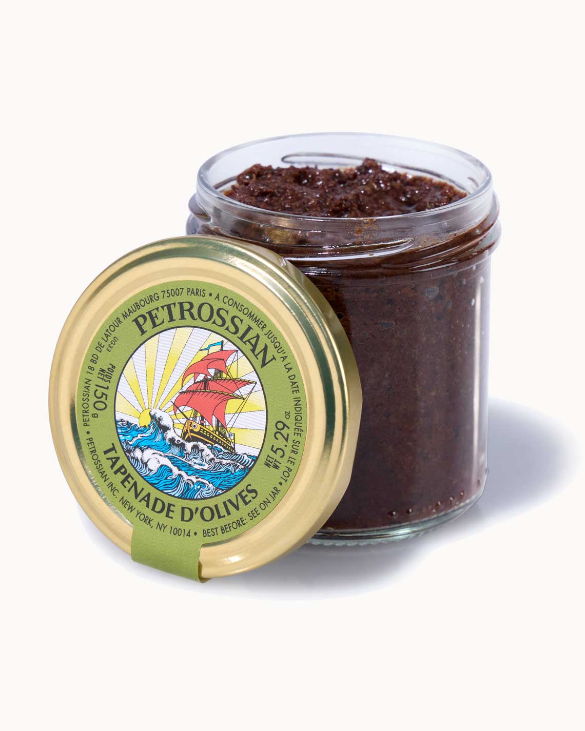 a short glass jar with a copper lid and olive green label sits on a white background. The jar is filled with dark green olive tapenade.