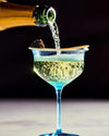 Champagne being poured into a blue-tinted coupe glass, with a wooden spoon holding black caviar on the rim. Effervescent bubbles rise against a dark background.