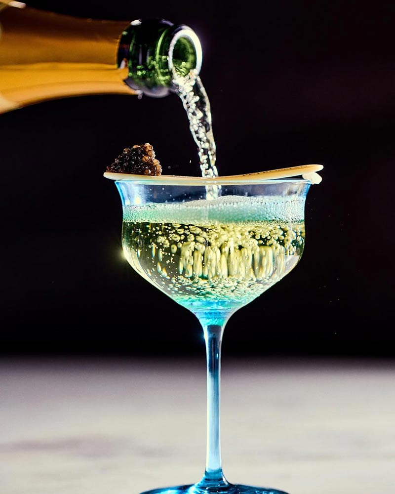 Champagne being poured into a blue-tinted coupe glass, with a wooden spoon holding black caviar on the rim. Effervescent bubbles rise against a dark background.