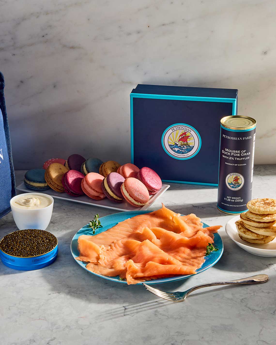 a collection of Petrossian delicacies arranged on a white marble counter, including a tin of caviar, a plate of smoked salmon, a tin of foie gras and french macarons.