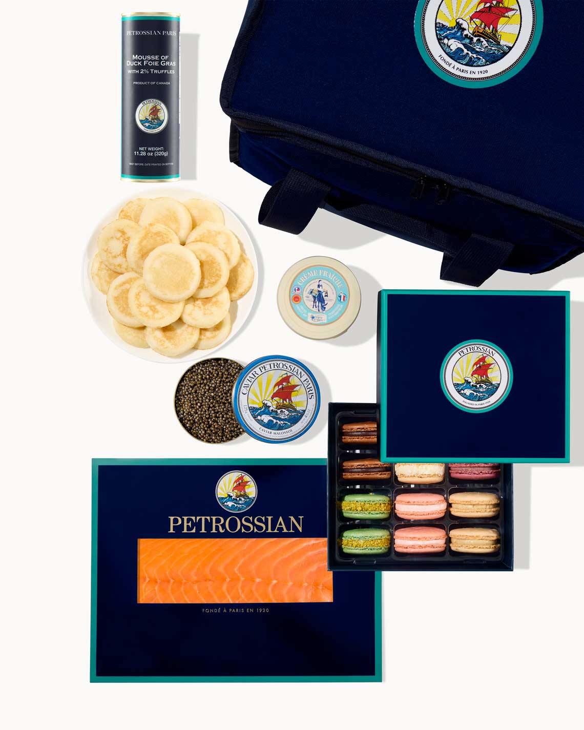 an overhead image of a navy-Blue Petrossian tote bag with blinis, caviar, french macrons, foie gras and smoked salmon laid out on a white background.