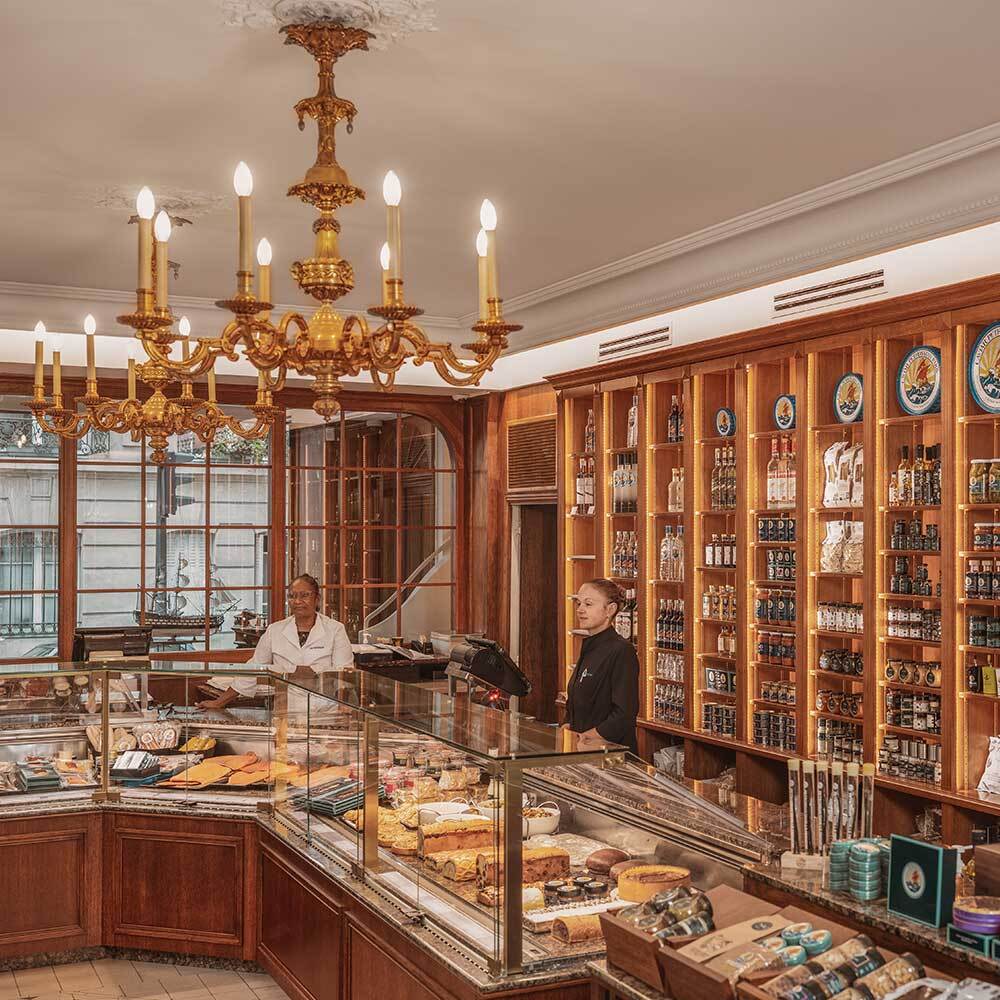 an interior image of the Petrossian Parisian boutique, in which two clerks stand among elegant wood shelves and glass counters. Many Petrossian products are on display.