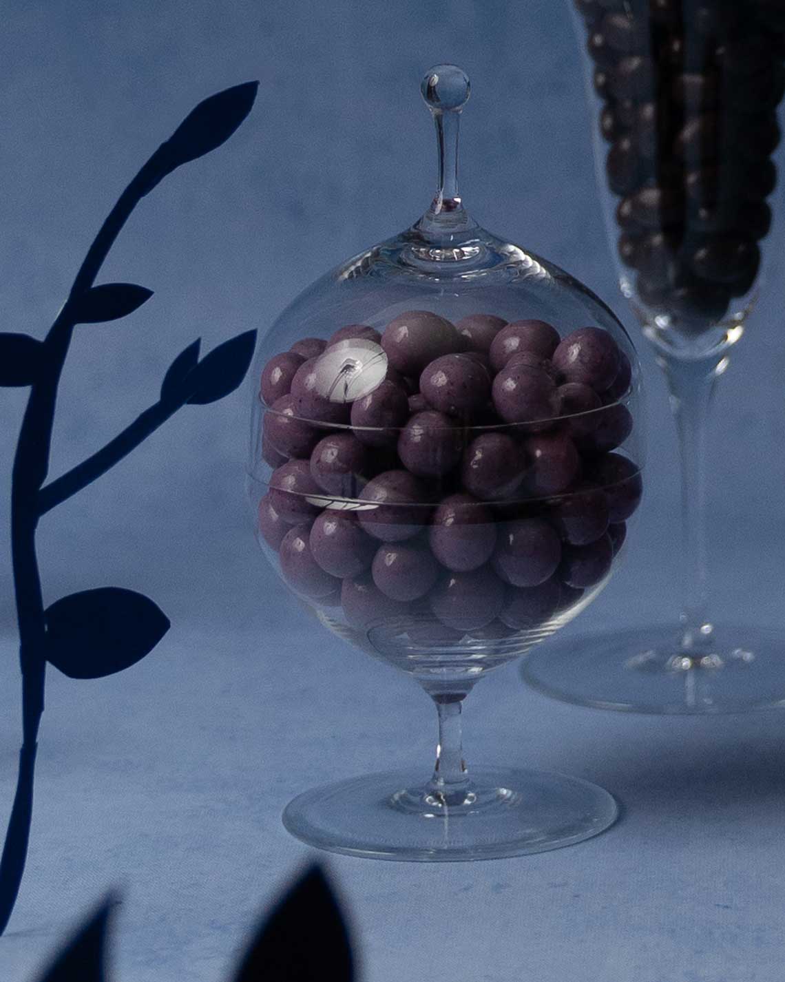 an elegant glass serving dish with a glass top sits on a textured deep blue background. Inside are two dozen large, purple chocolate covered blueberries