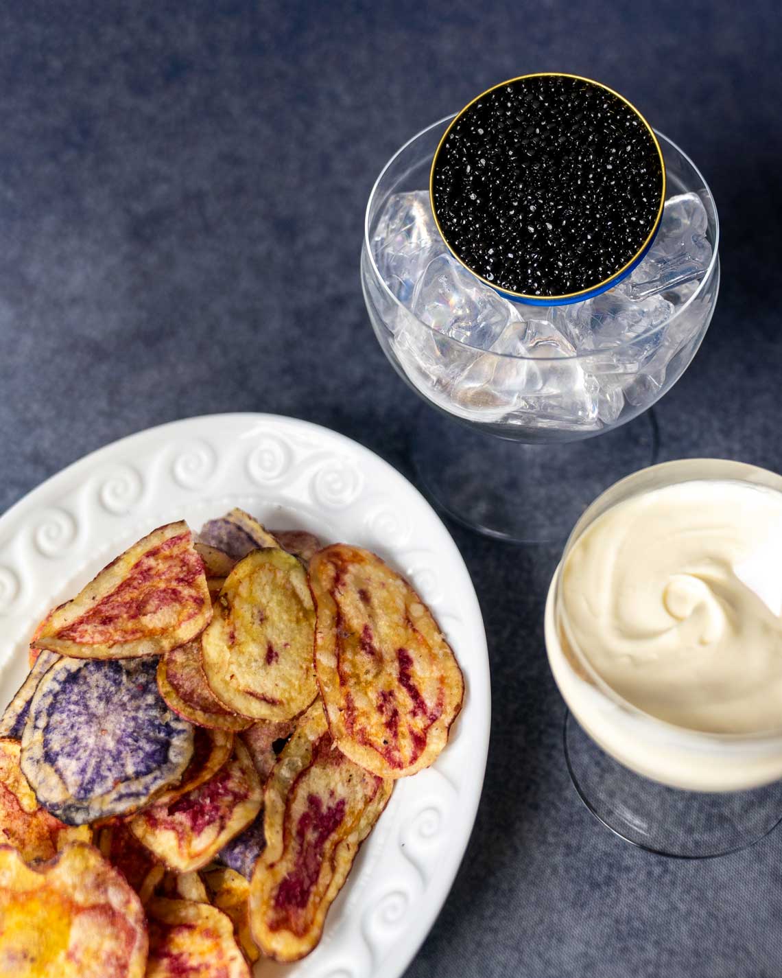 a top-down image of a plate of gorgeous, multi-colored potato chips on a white plate. Next to the plate is a glass of creme fraiche and a glass filed with ice and a tin of black baika caviar.