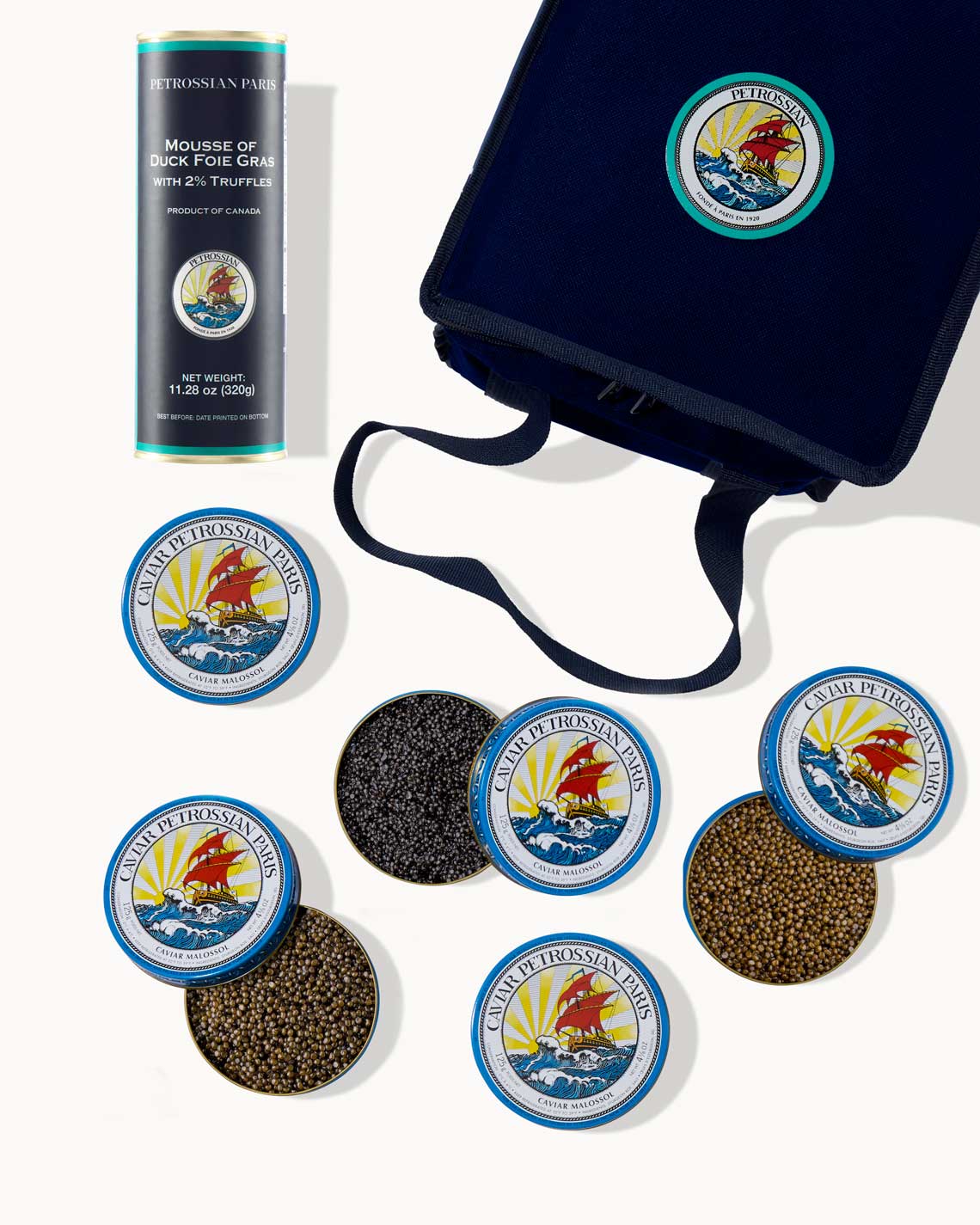an overhead image of a navy-Blue Petrossian tote bag with five tins of different caviars and a canister of foie gras mousse laid out on a white background.