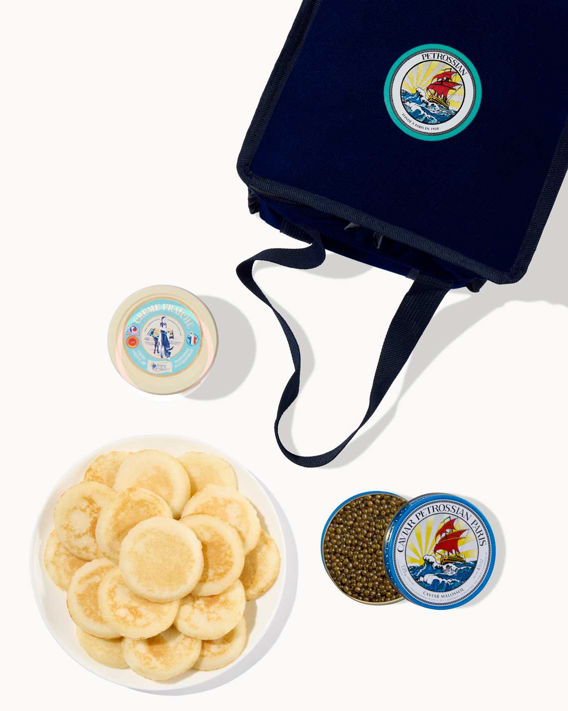 an overhead image of a navy-Blue Petrossian tote bag with a tin of caviar, a plate of blinis and a jar of creme fraiche laid out on a white background.