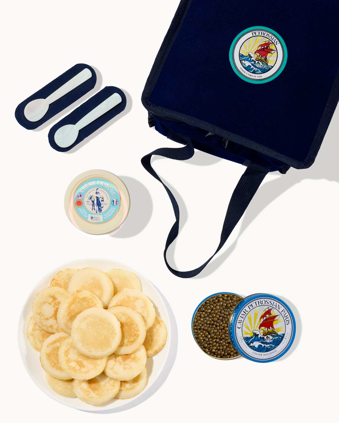 an overhead image of a navy-Blue Petrossian tote bag with a tin of caviar, a plate of blinis and a jar of creme fraiche laid out on a white background.