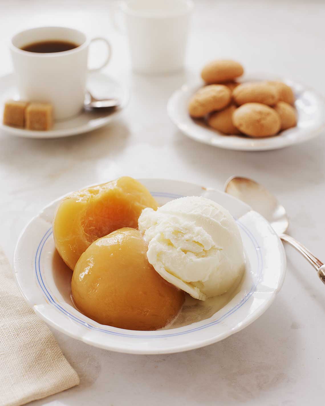 Two luscious peach halves sit in a white bowl with a generous scoop of vanilla ice cream. There are coffee and gingersnap cookies in the background.