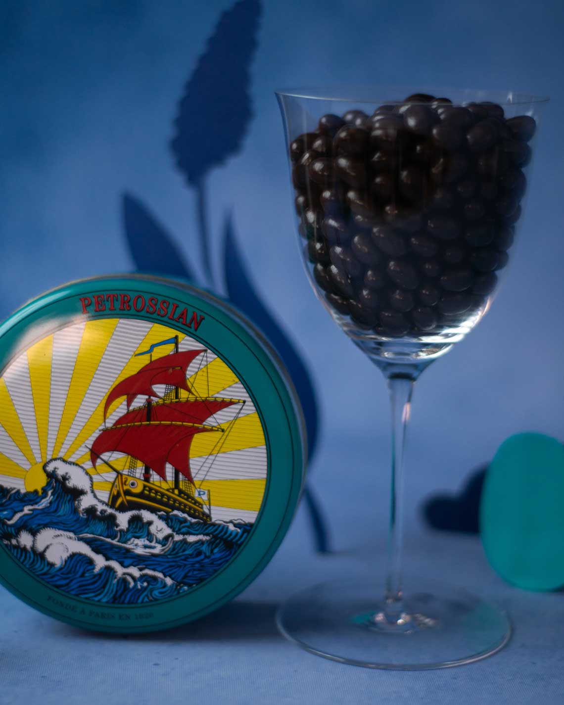 an elegant glass filled with black, vodka chocolates sits next to a green chocolate tin on a blue background with colorful paper cut-outs of plants in the background.