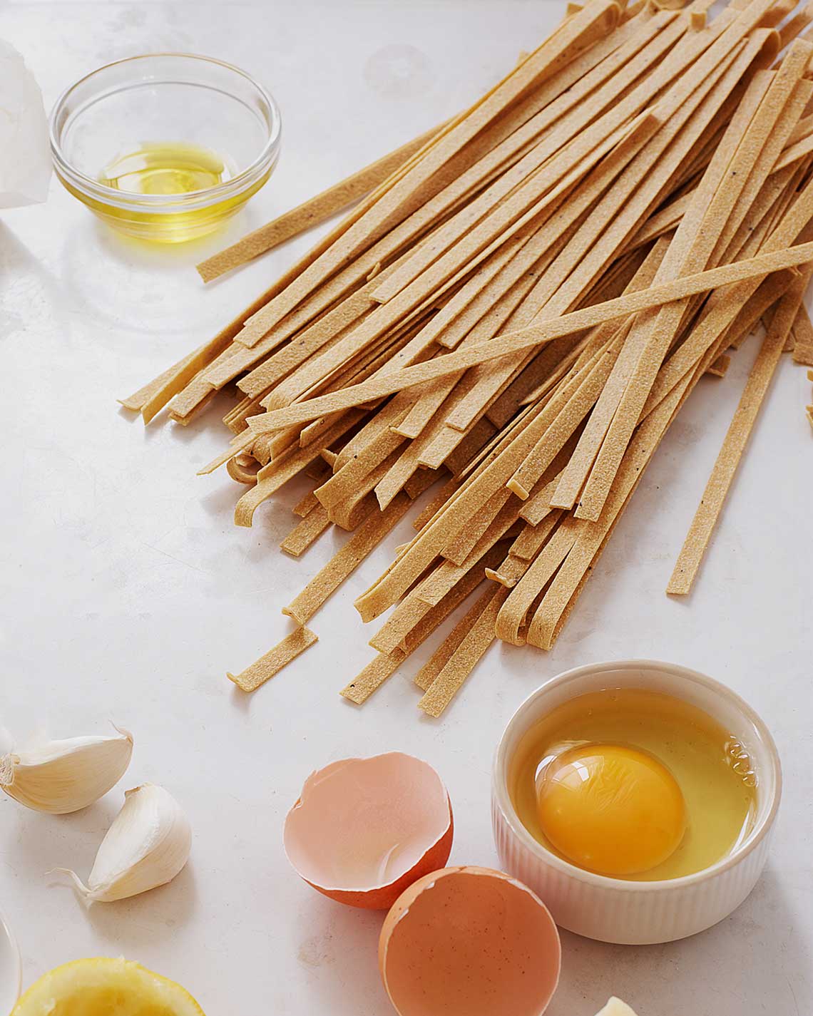 a light brown fettucine is laid out, still uncooked, on a white countertop next to egg shells and bowl with a raw egg sits, ready for prep.