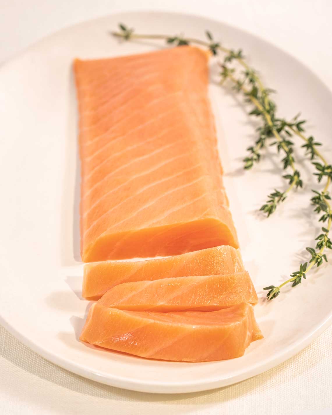 a long, thin filet of light orange smoked salmon rests on an off-white plate on a beige tablecloth. the tick slices of salmon are garnish with rosemary.