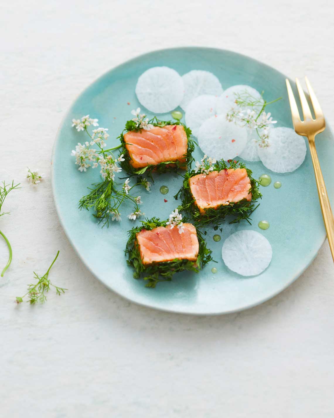 three luscious slices of smoked salmon sit on a teal blue plate, with a golden fork garnished with watercress, herbs and white flowers.