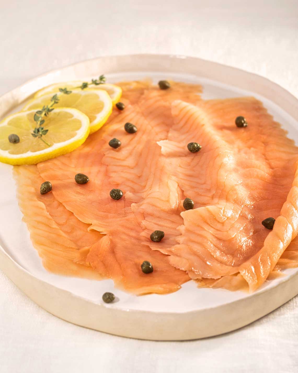 lush slices of pale orange smoked salmon are plated lavishly on a white platter with capers and lemons for a garnish