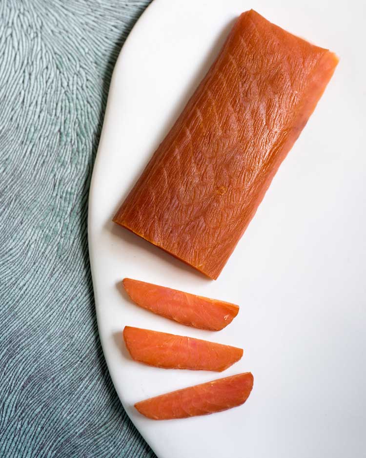 a firm, hearty cut of Souhoi smoked salmon lies, partially sliced on a white plate on a grey, patterned background.