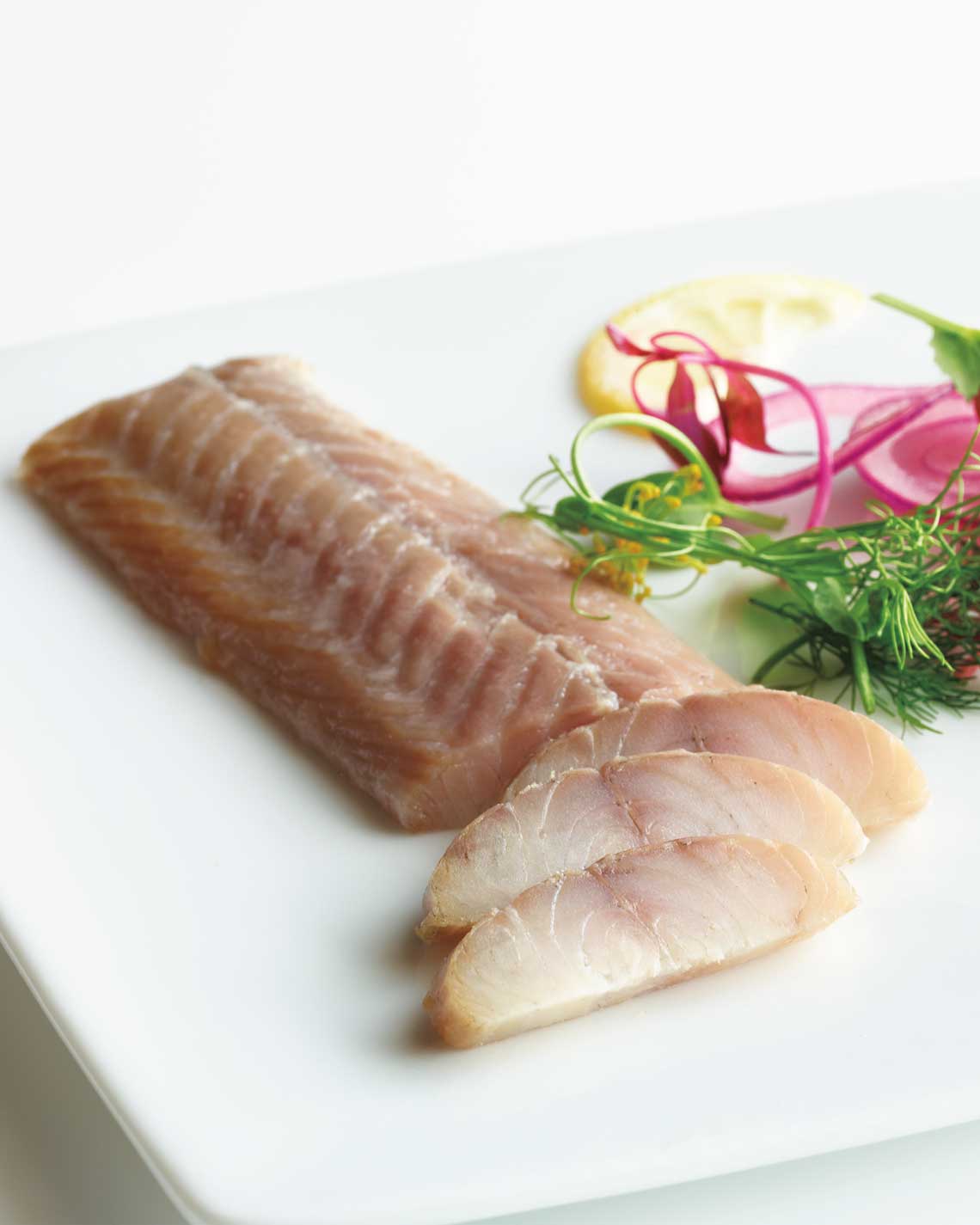 a thin filet of light grey smoked eel is plated and partially sliced on a white porcelain platter with a garnish of greenery, lemons and red onions. 