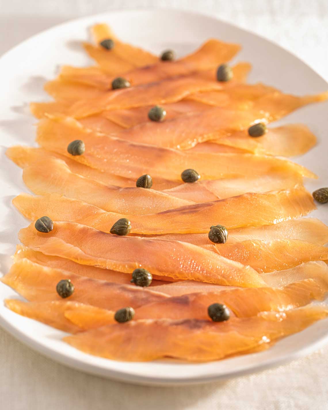 light, tempting slices of cold-smoked trout lie in a cascading pattern on a long white plate, garnished with capers. 