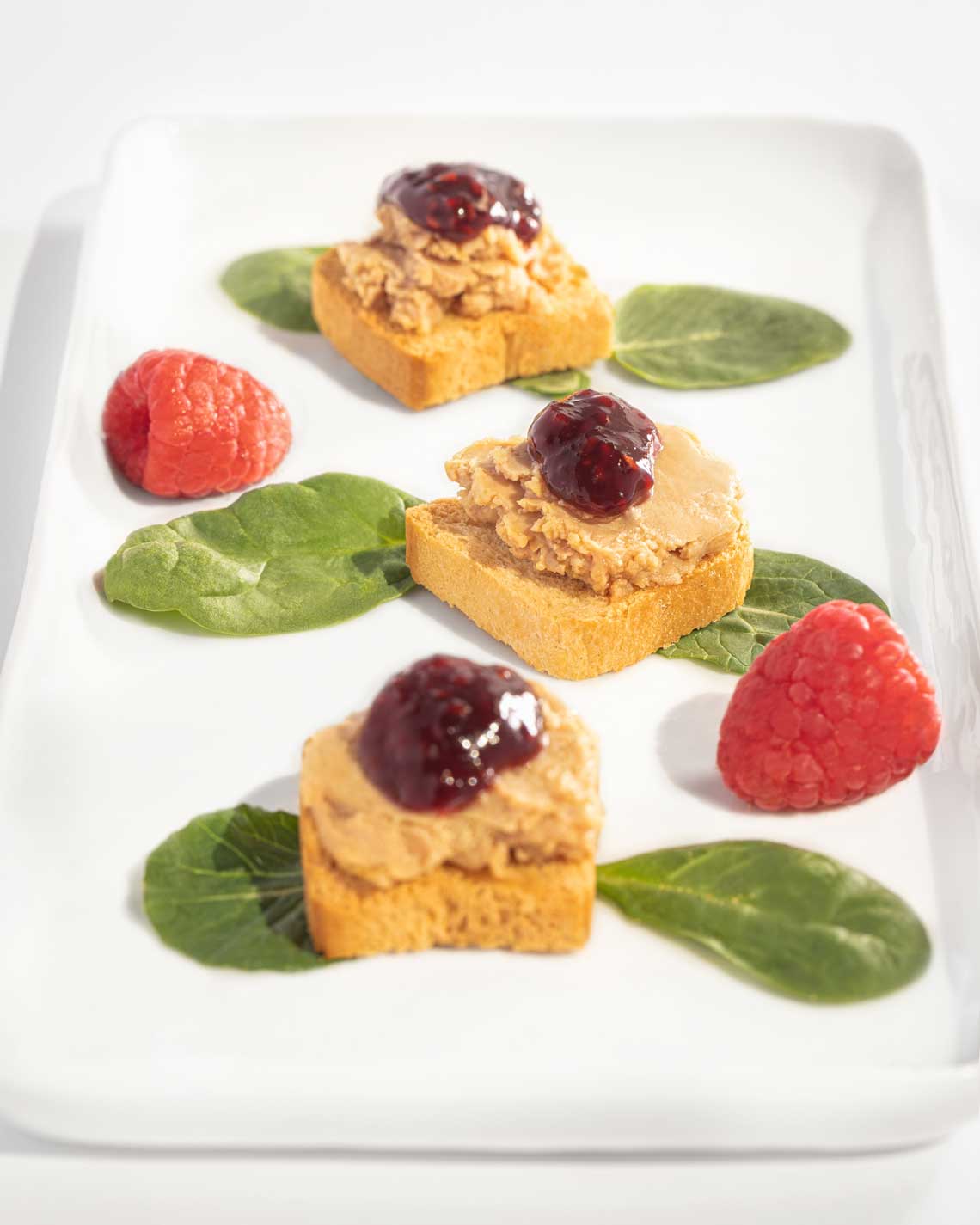 three light portions of foie grs are plated on baguette mini-toasts with dollops of raspberry jam on top and raspberries for garnish. 
