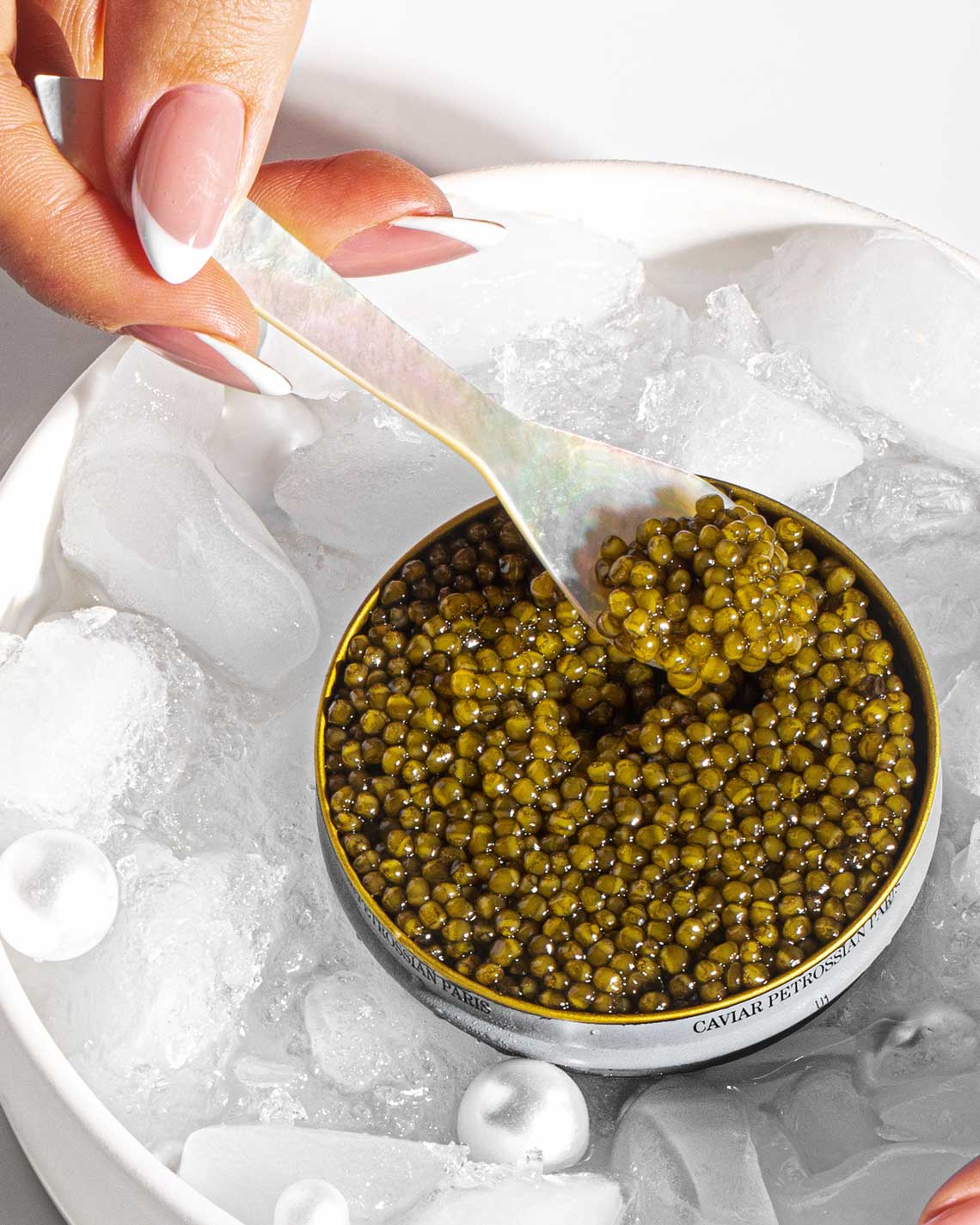 A woman's hand holds an elegant pearl spoon in an open tin of moist, bright green Kaluga-Hybrid caviar. The tin is sitting in a bowl of ice.
