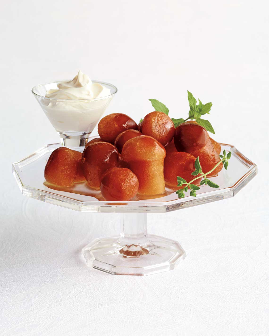 a raised glass serving plate covered in syrupy plump babas au rhum, plated with herbs and a glass or thick cream
