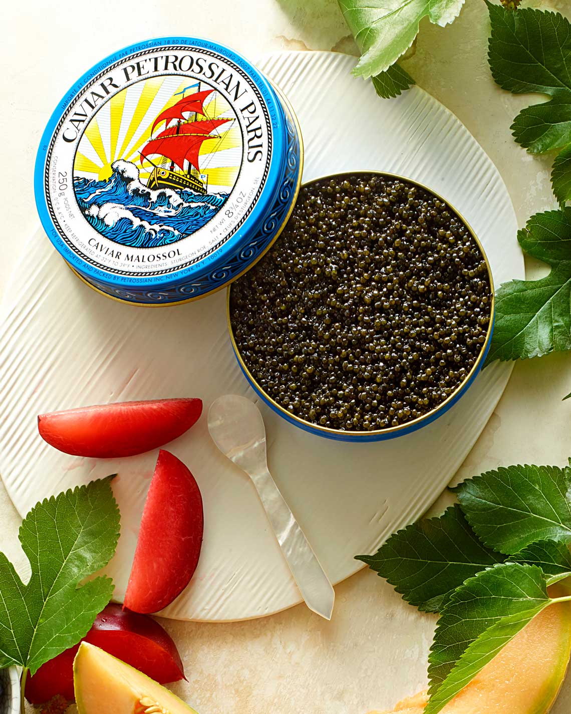 an open Petrossian tine of dark green caviar sits on a white ceramic platter surrounded by vibrant greenery and bright red and yellow fruit slices