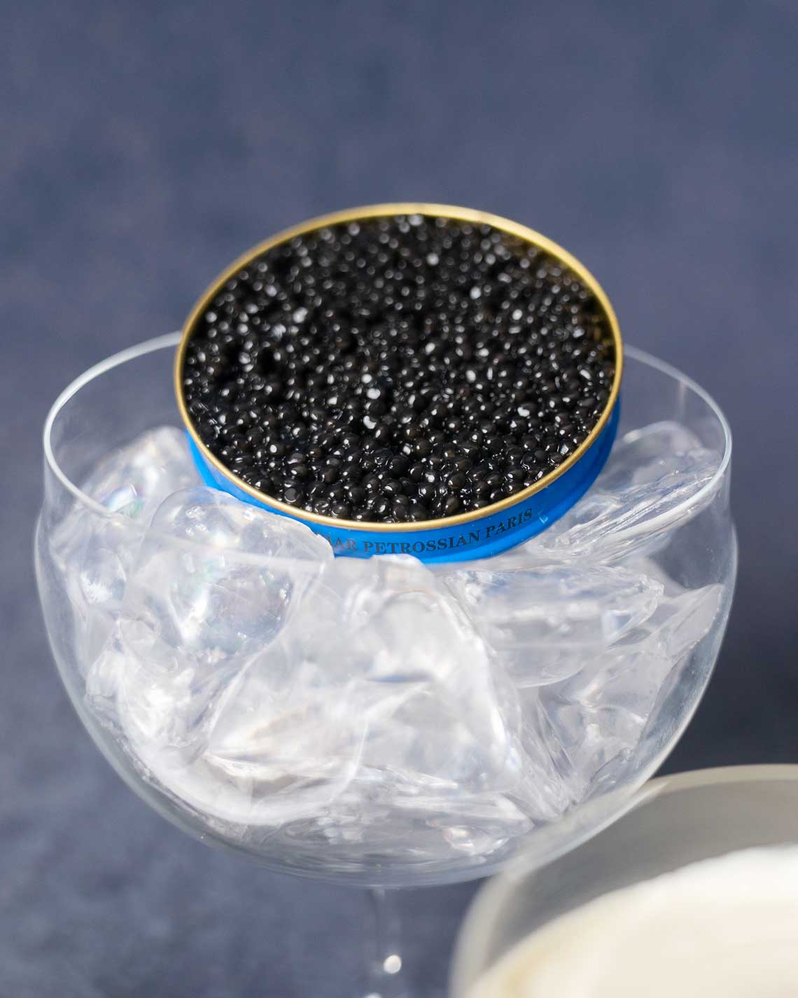 a close-up of a glass, filled with ice and holding up a blue Petrossian caviar tin, filled with black, tempting baika caviar on a navy blue background.