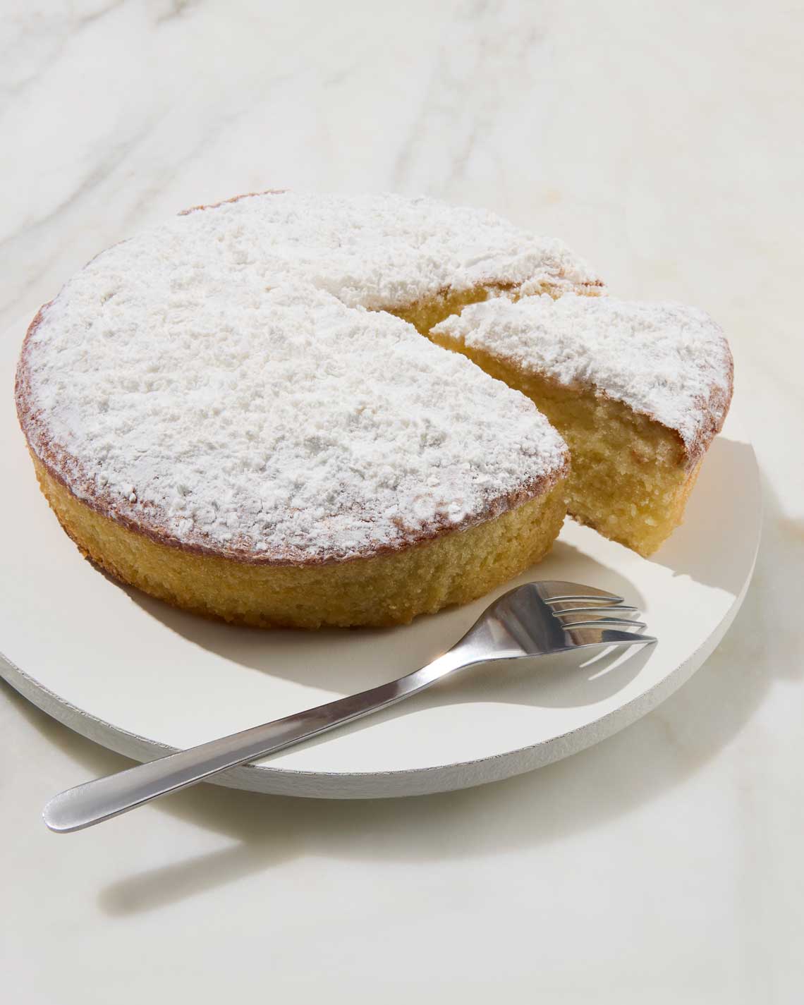 a yellow bourbon cake with a powdered top on a white plate with a single slice peeking out and a silver fork