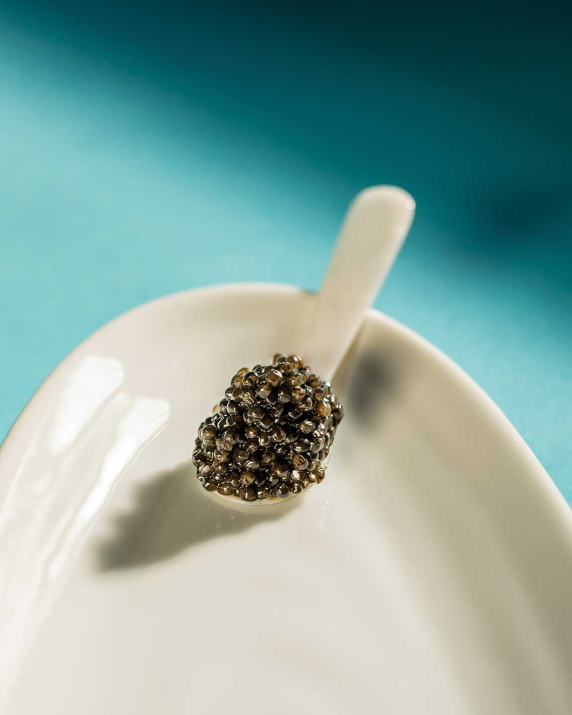 a white pearl spoon filled with a generous dollop of caviar sits on the edge of a polished china plate.