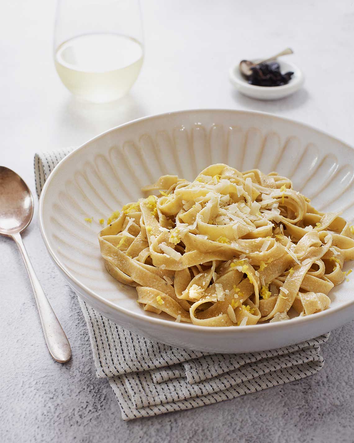  a rich bowl of  light green truffled tagliatelle sits on a clothe napkin on a white countertop. a glass of white wine and a metal spoon sit nearby/