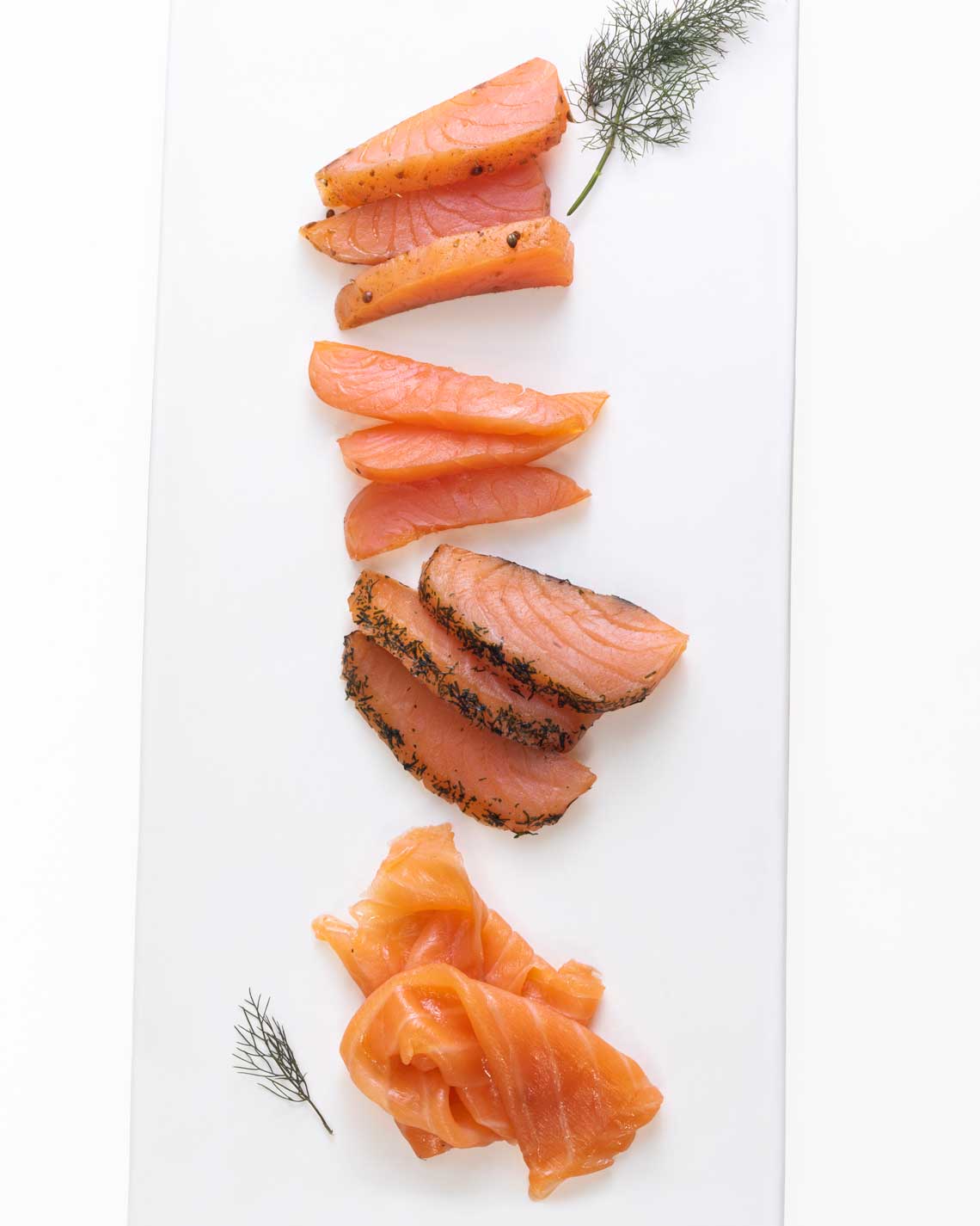 four types of smoked salmon are stylishly arranged on a white cutting board with sprigs of dill as a garnish